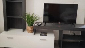 A modern living room with a flat-screen TV on a dark wooden stand, a white cabinet with drawers, and a woven plant on the cabinet. - Apartment and Room Mare Vrlika - Double Bedroom with Terrace and External Kitchenette (Sinj)