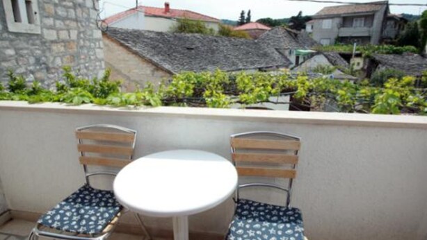 A balcony with two metal chairs and a small round table, overlooking a residential area with stone buildings and greenery.