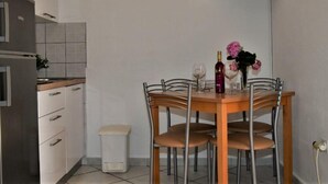 A compact kitchen with white cabinets, a small trash bin, and a dining table with four chairs. A flat-screen TV is mounted on the wall above the table.