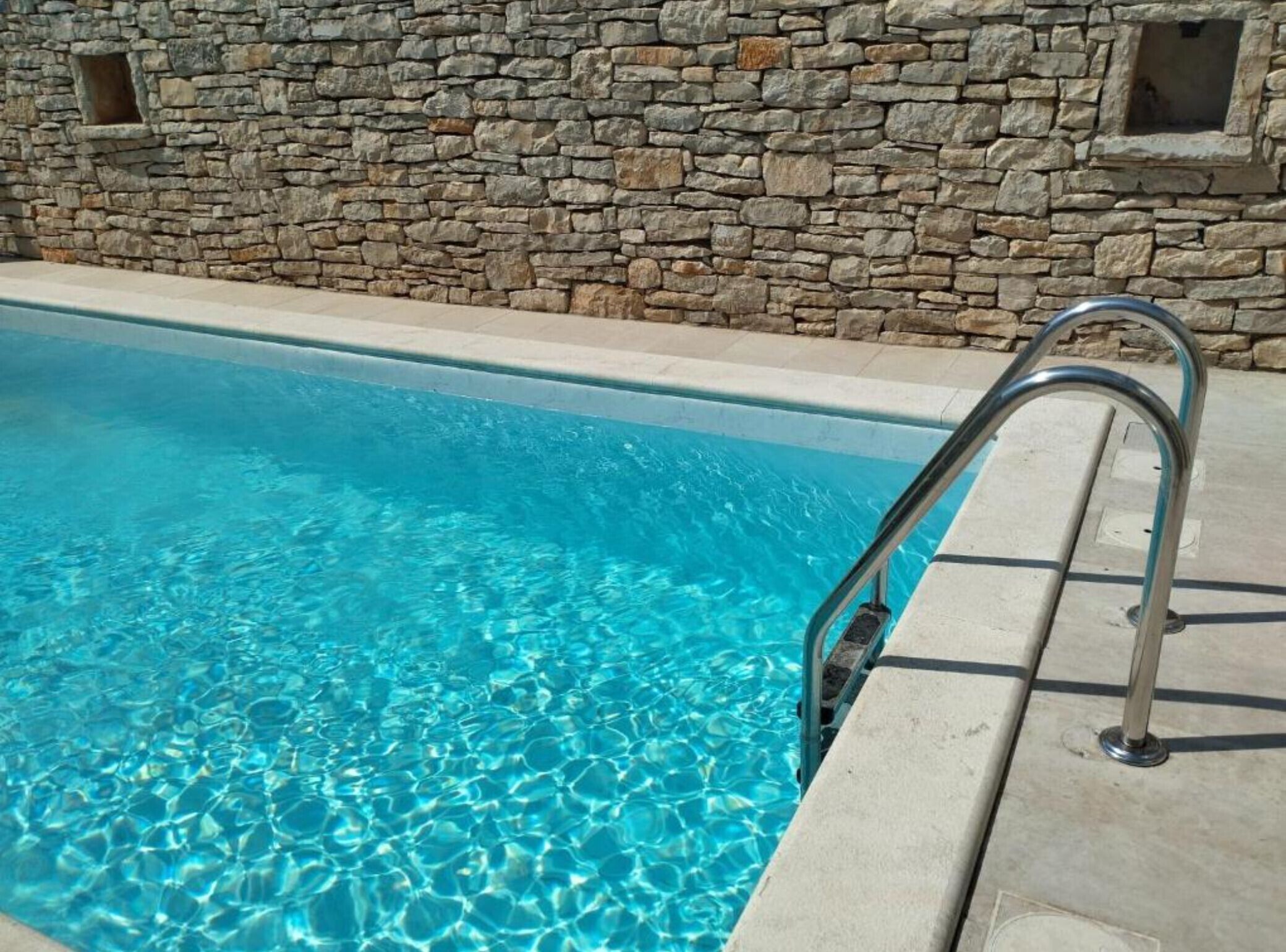 Une piscine à l’eau d’un bleu cristallin, un mur en pierre et un escalier métallique pour y accéder.