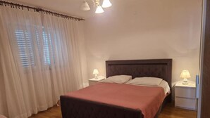 A bedroom with a large bed, two bedside tables with lamps, a window with blinds, and a wooden floor. - Apartments Bilić Trogir - Two-bedroom Apartment With Sea View (A1) (Trogir)