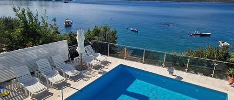 A pool with clear blue water, surrounded by lounge chairs and umbrellas, overlooking a calm sea with boats.