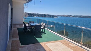 A balcony with a table and chairs, overlooking a body of water and a distant shoreline. - Apartments Bilić Trogir - Three-Bedroom Apartment With Sea View (A2) (Trogir)
