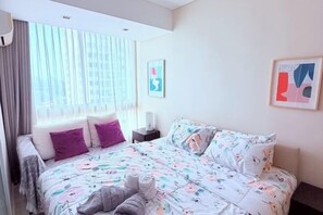 Una habitación de hotel con cama, almohadas, una mesita de noche, una ventana con cortinas y dos cuadros en la pared.