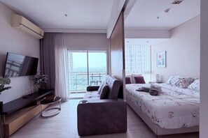 Habitación de hotel moderna con televisor de pantalla plana, sofá, cama con ropa de cama floral, balcón con vistas y aire acondicionado instalado.