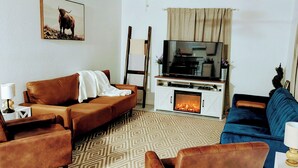 Een woonkamer met een bruine bank, een blauwe bank, een open haard, een televisie en een schilderij van een koe aan de muur.