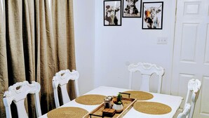 Een eettafel met een witte tafelkleed, omringd door witte stoelen met decoratieve rugleuningen. Daarboven hangen drie ingekaderde foto’s aan de muur.