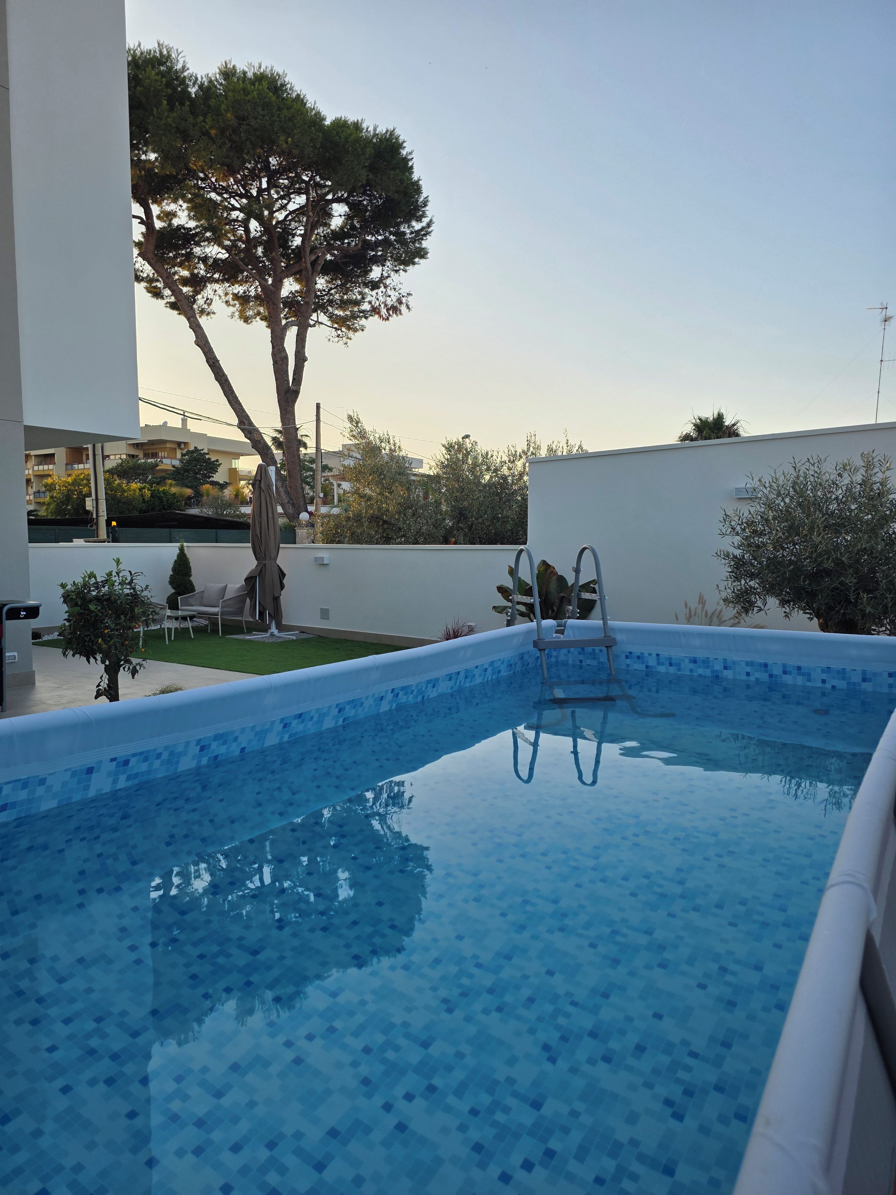 A swimming pool with a tiled edge, a ladder, and a small deck area with chairs.