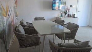 A modern dining area with a glass table and wicker chairs, a flat-screen TV mounted on the wall, and a white door in the background. - M4ree Seaside Suite And Relax (Bari)