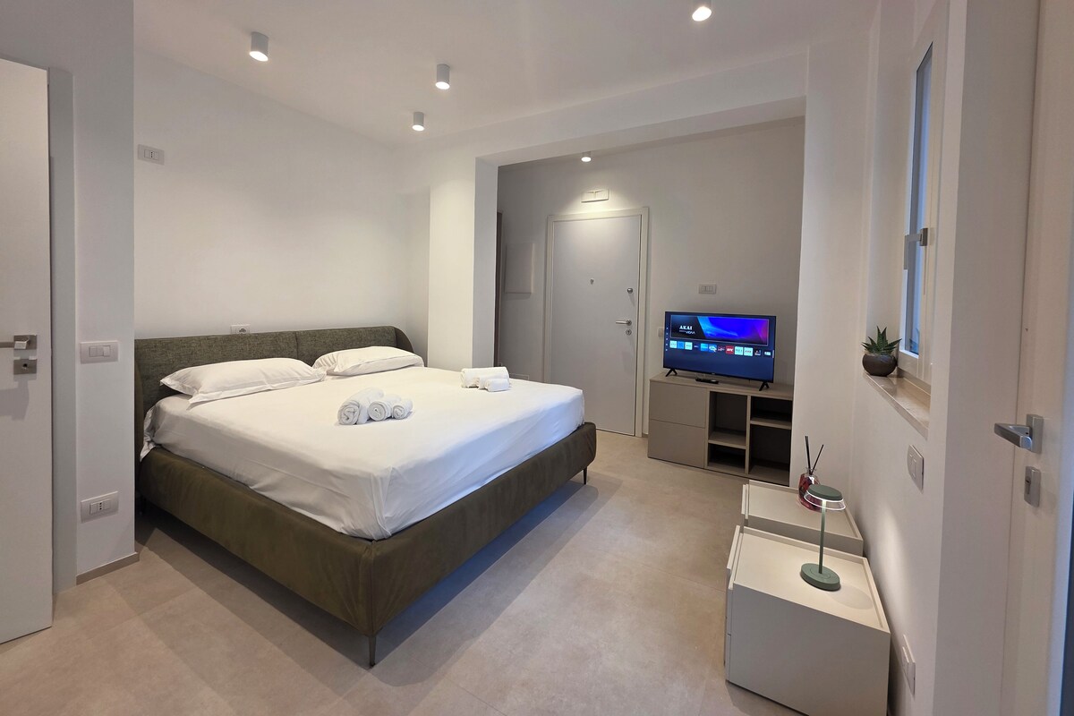 A modern hotel room with a large bed, a flat-screen TV, a small desk, and a potted plant.