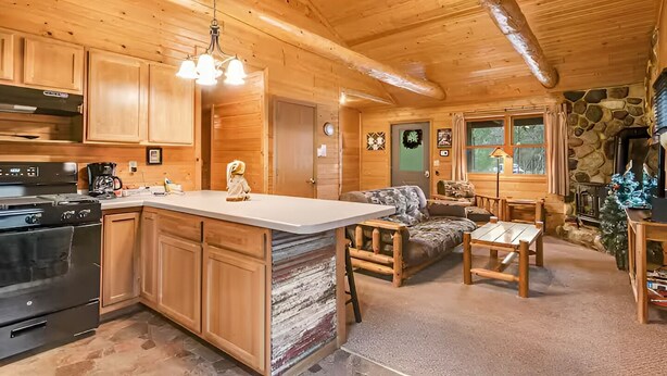 Un intérieur de chalet chaleureux avec des murs en bois, une cheminée en pierre, un coin repas et une cuisine dotée d’un grand îlot central.