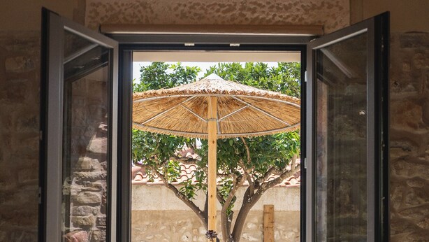 Een kamer met een bed, een glazen deur naar een buiten zitgedeelte met rieten stoelen en een rieten parasol.