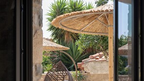 Een kamer met een glazen deur die uitkomt op een terras met een rieten stoel, een tafel en een rieten parasol.