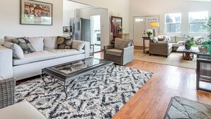 A spacious living room with a sofa, coffee table, and wicker chairs. A kitchen is visible in the background. - 2 Mi to Downtown Coos Bay: Home w/ Deck, Fire Pits (Coos Bay)