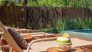 Zona junto a la piscina con sillones de mimbre, una mesa de madera con un cuenco y una rodaja de limón, y una valla de madera con la palabra "KOKOYE" grabada.