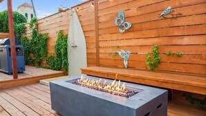 A concrete fire pit with flames, surrounded by wooden decking and a wooden wall with decorative metal birds and butterflies.