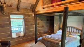 Une chambre rustique aménagée dans un chalet en rondins, avec un lit à baldaquin, un banc, un fauteuil en cuir et une fenêtre donnant sur un paysage verdoyant.