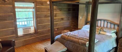 A rustic log cabin room with a four-poster bed, a bench, a leather chair, and a window with a view of greenery.