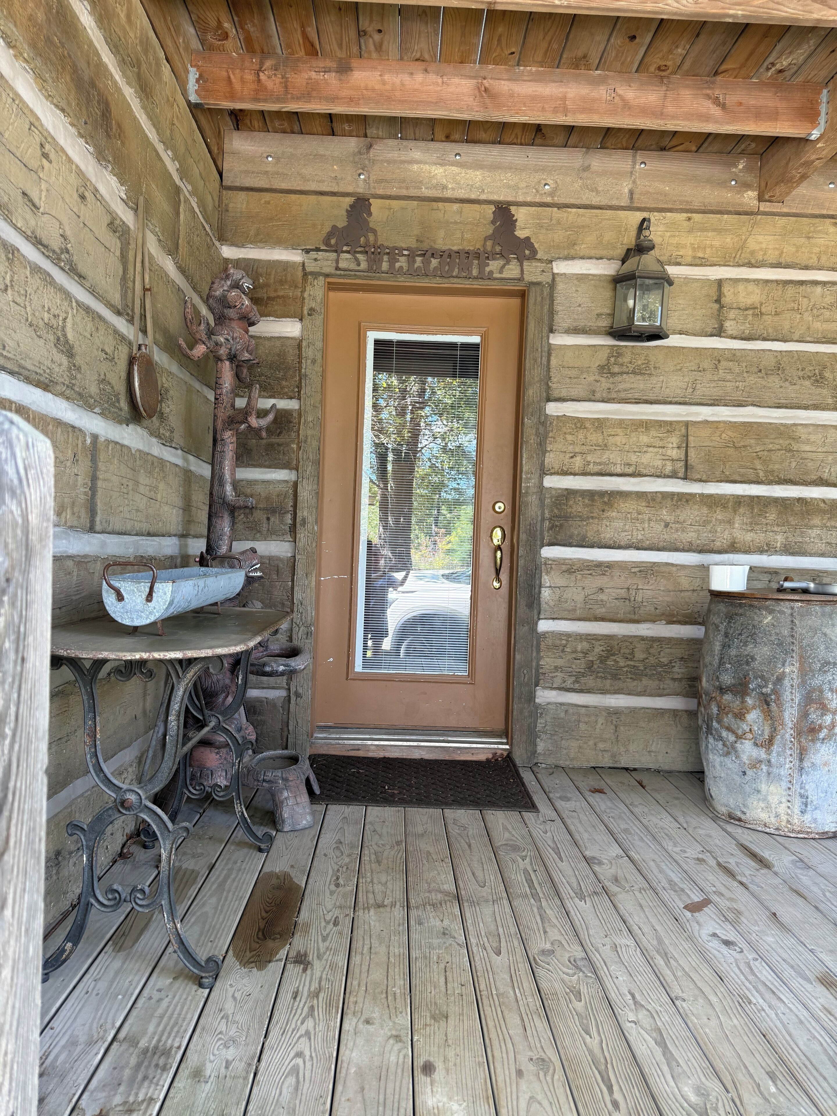 A wooden cabin entrance with a brown door, a welcome sign, a lantern, and a rustic metal table.
