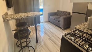 A compact kitchen with a gas stove, a small bar stool, a grey sofa, and a ceiling light fixture. - Charming 1-bedroom studio facing the pool, in the heart of Sosua (Sosúa)