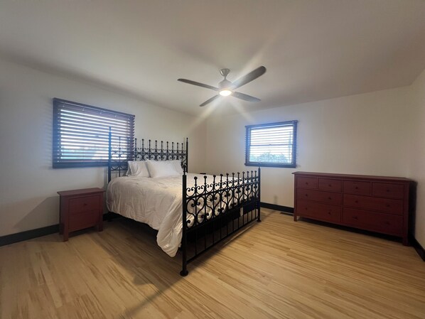 A bedroom with a bed, a ceiling fan, two windows with blinds, a dresser, and a nightstand.