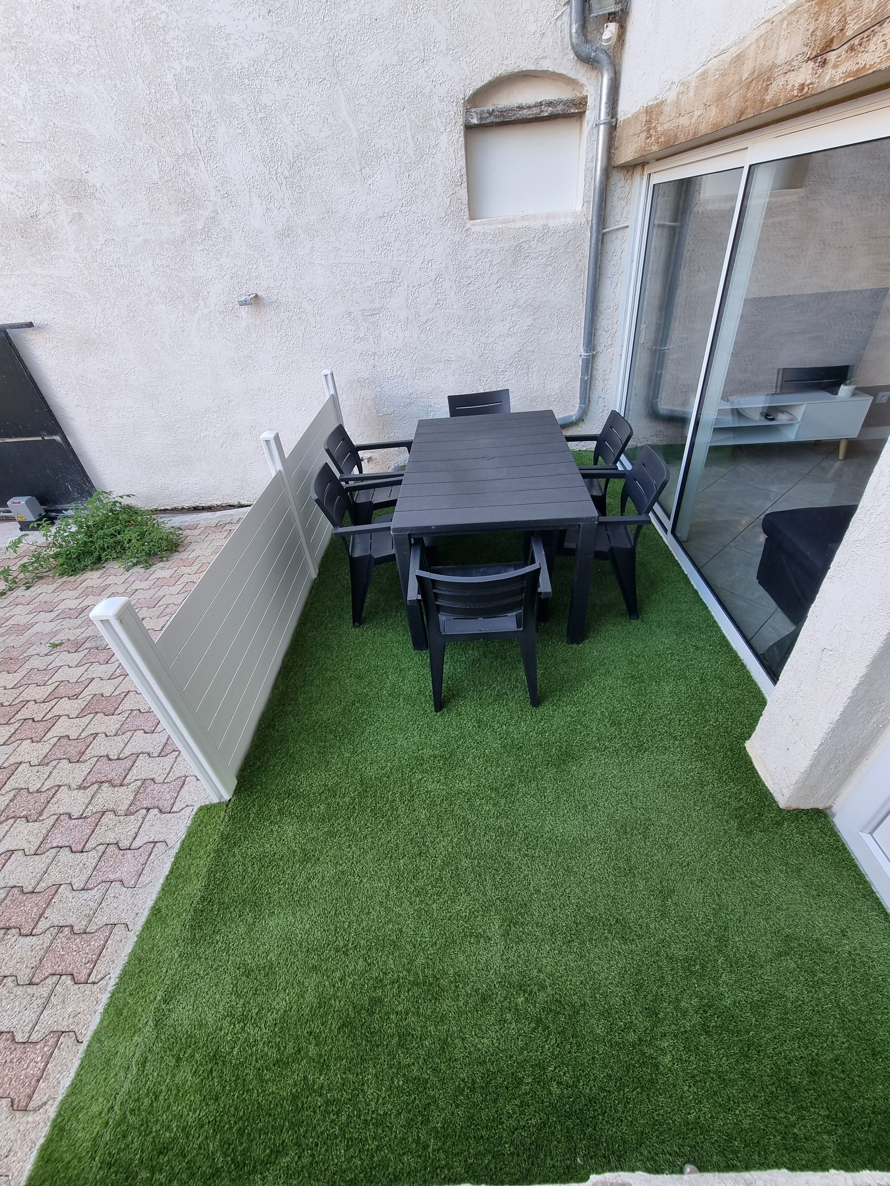 A patio with a table and chairs, artificial grass, a white wall, and a sliding glass door.
