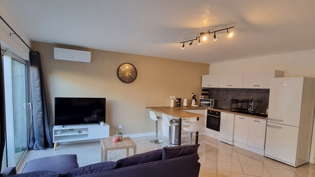 Een moderne woonkamer met een donkergrijze bank, een flatscreen televisie en een eethoek met een houten tafel en witte stoelen. De keuken heeft witte kastjes, een koelkast en een magnetron.