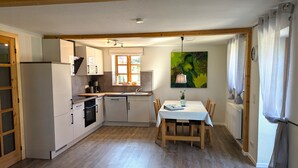 A compact kitchen with white cabinets, a built-in oven, and a dishwasher. There's a dining table with chairs, a window with curtains, and a green painting on the wall. - Koppelkuck vacation apartment on a beautiful vacation farm with pool and sauna Ostseena (Schwartbuck)