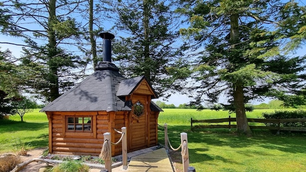A wooden cabin with a chimney, surrounded by a wooden deck and a rope barrier. - Koppelkuck vacation apartment on a beautiful vacation farm with pool and sauna Ostseena (Schwartbuck)