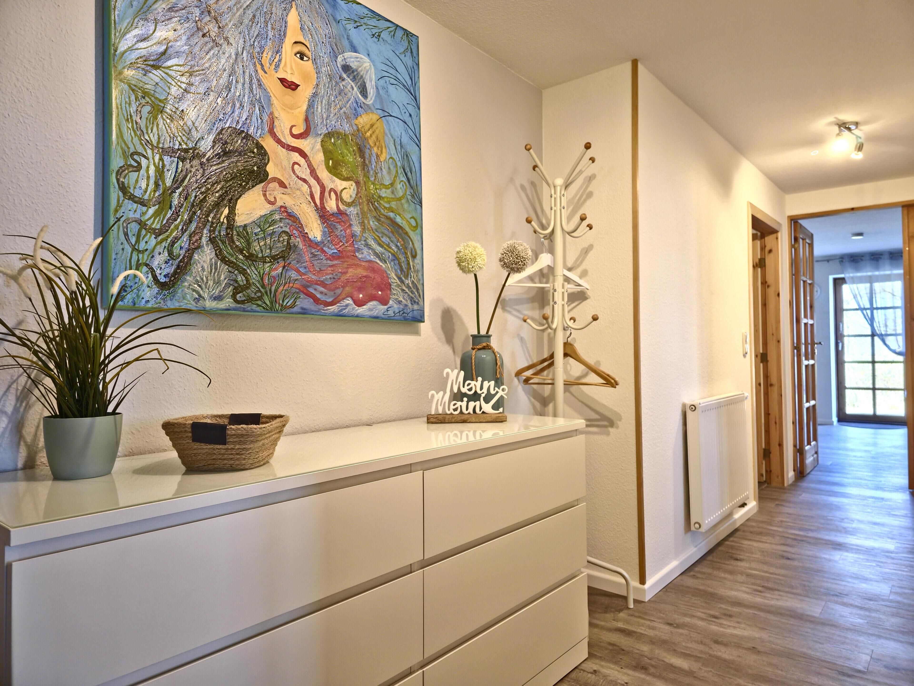 A hallway with a white console table, a painting of a mermaid, a coat rack, and a plant.