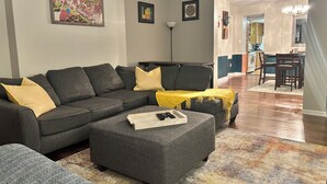 A living room with a sectional sofa, ottoman, and a colorful rug. There is a dining area visible in the background. - Cozy, spacious 1 bdrm apt in Bloomfield with off street parking. Great location! (Pittsburgh)