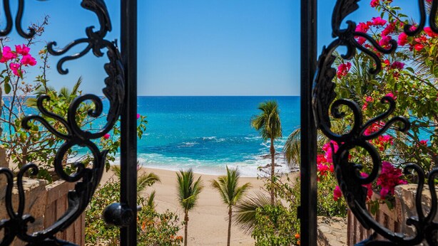 Blick auf einen Strand durch kunstvolle Metallgitter mit rosa Blumen und Palmen.