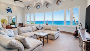 Ein geräumiges Wohnzimmer mit großem Fenster zum Meer, einem Deckenventilator und einer an der Wand lehnt Gitarre.
