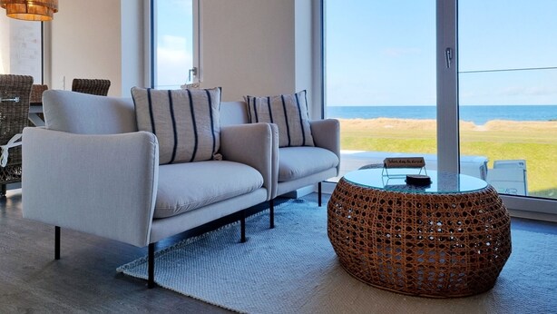 A modern living room with a sofa, a round wicker coffee table, and a large window offering a view of the beach. - Baltic Sea vacation apartment Surfside - sea view from every window (Schönberg (Holstein))