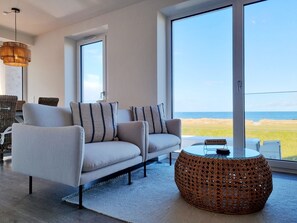 A modern living room with a sofa, a round wicker coffee table, and a large window offering a view of the beach. - Baltic Sea vacation apartment Surfside - sea view from every window (Schönberg (Holstein))
