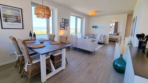 A modern living room with a sofa, dining table with wicker chairs, and a view of the sea through large windows. - Baltic Sea vacation apartment Surfside - sea view from every window (Schönberg (Holstein))