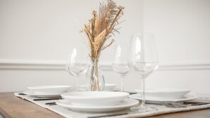 A dining table set with white plates, clear wine glasses, and a vase with dried flowers.