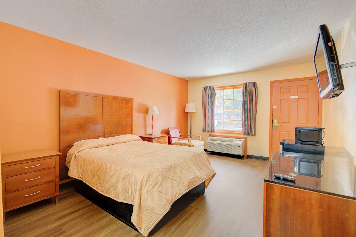Une chambre d’hôtel avec un lit en bois, un bureau avec une télévision, une chaise, une table de chevet et une fenêtre avec des rideaux.