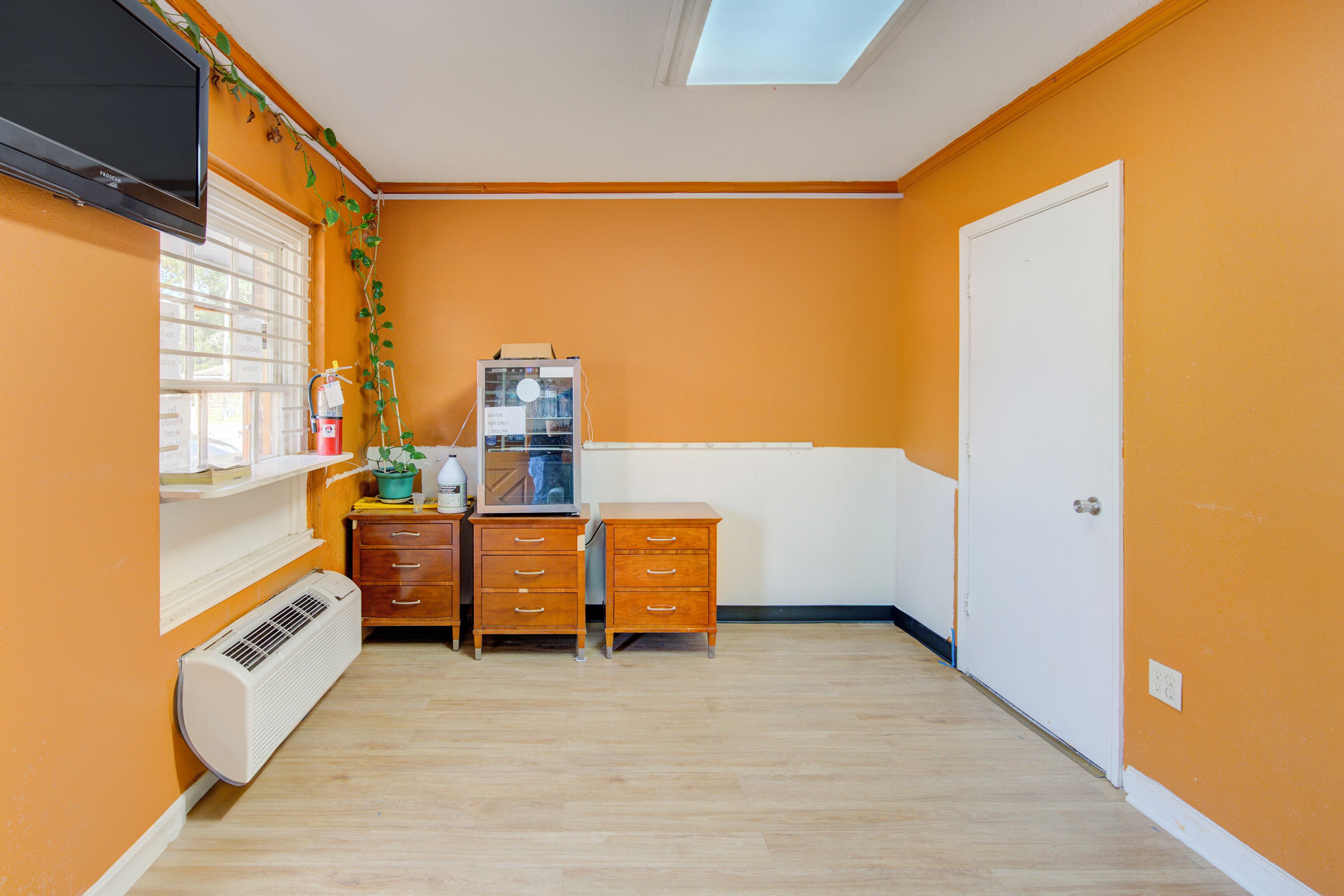 Habitació amb parets taronja, una porta blanca, una finestra amb persianes, un televisor fixat a la paret i un escriptori petit amb calaixos.