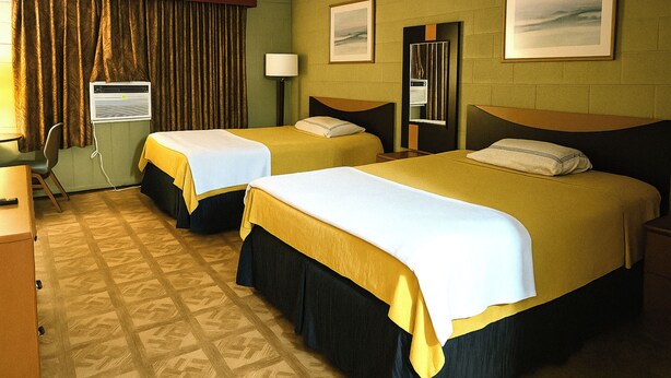 Ett hotellrum med två sängar, ett skrivbord, en stol, en luftkonditioneringsanläggning och två inramade tavlor på väggen.