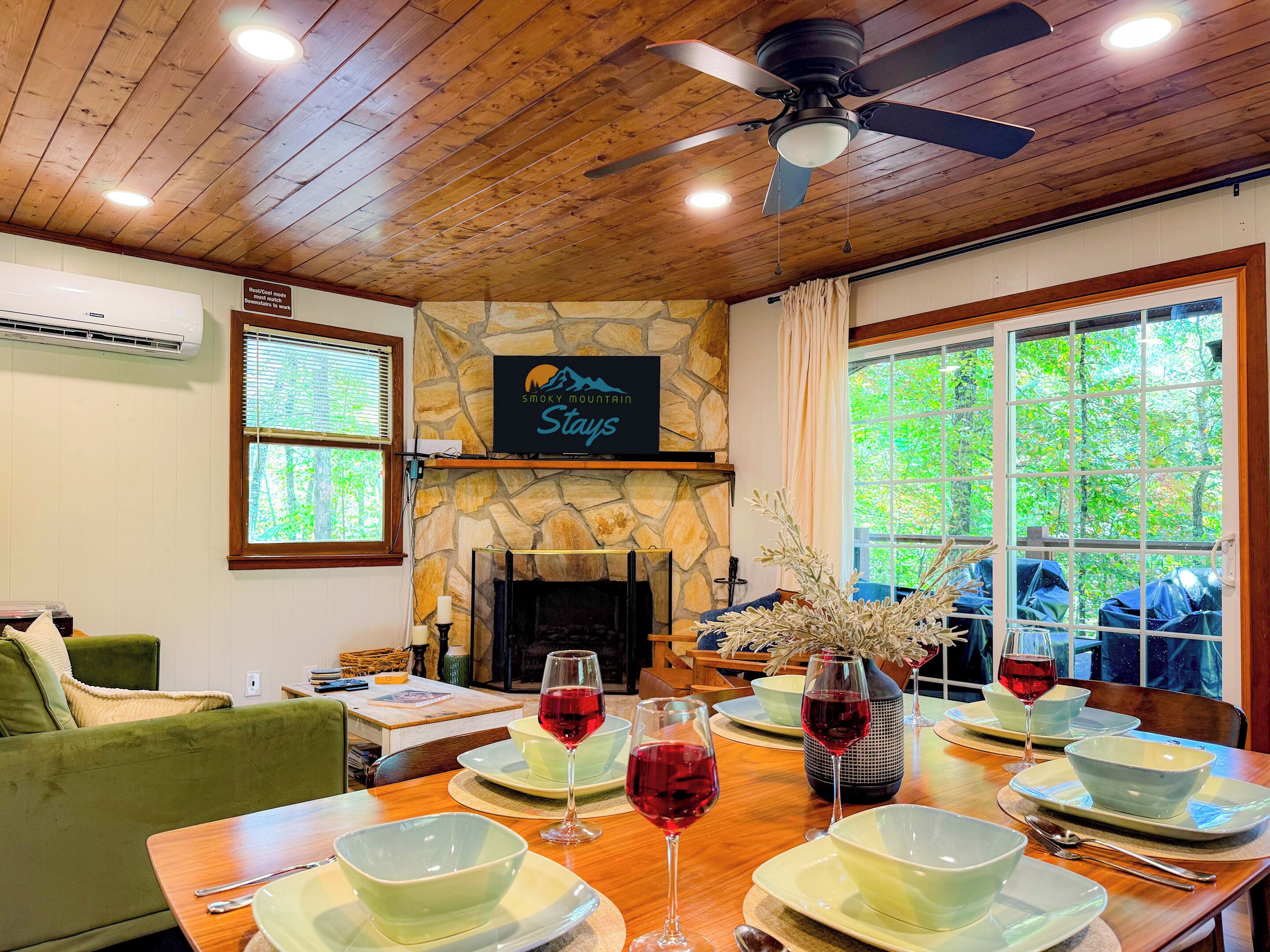 A dining area with a stone fireplace, a table set for four with wine glasses, and a sign above the fireplace reading "Smoky Mountain Stays".