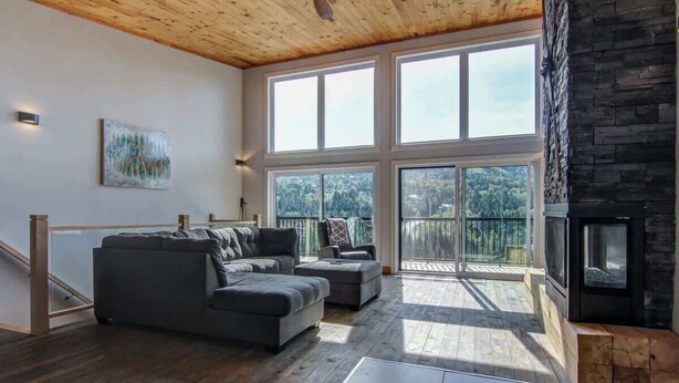 Un salon spacieux avec un grand canapé d’angle, une cheminée et un balcon offrant une vue sur les arbres.