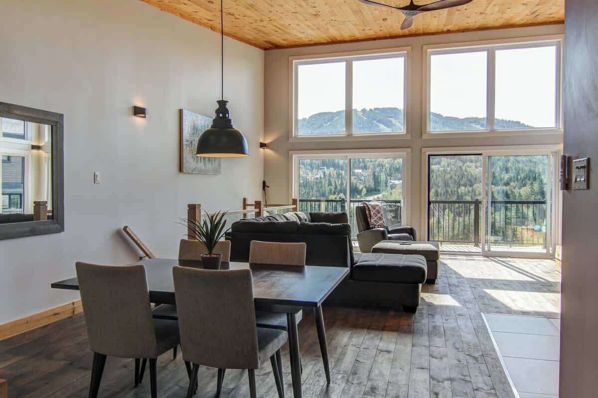 Un vaste espace de vie doté d’un plafond en bois, de grandes fenêtres offrant une vue sur les montagnes, d’une table à manger avec des chaises et d’un canapé.