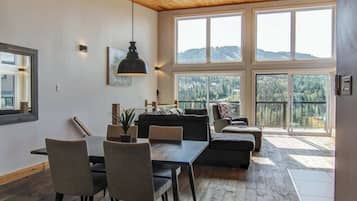 Un vaste espace de vie doté d’un plafond en bois, de grandes fenêtres offrant une vue sur les montagnes, d’une table à manger avec des chaises et d’un canapé.