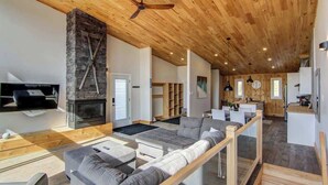 Un vaste espace de vie agrémenté d’une cheminée en pierre, d’un plafond en bois et d’une cuisine moderne en arrière-plan.
