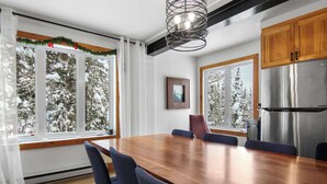 Un espace repas moderne avec une table en bois et des chaises bleues, de grandes fenêtres donnant sur des arbres couverts de neige, et un réfrigérateur en acier inoxydable.