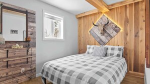 Une chambre à coucher avec une tête de lit en bois, une couvre-lit à carreaux, un miroir et une fenêtre donnant sur des arbres.