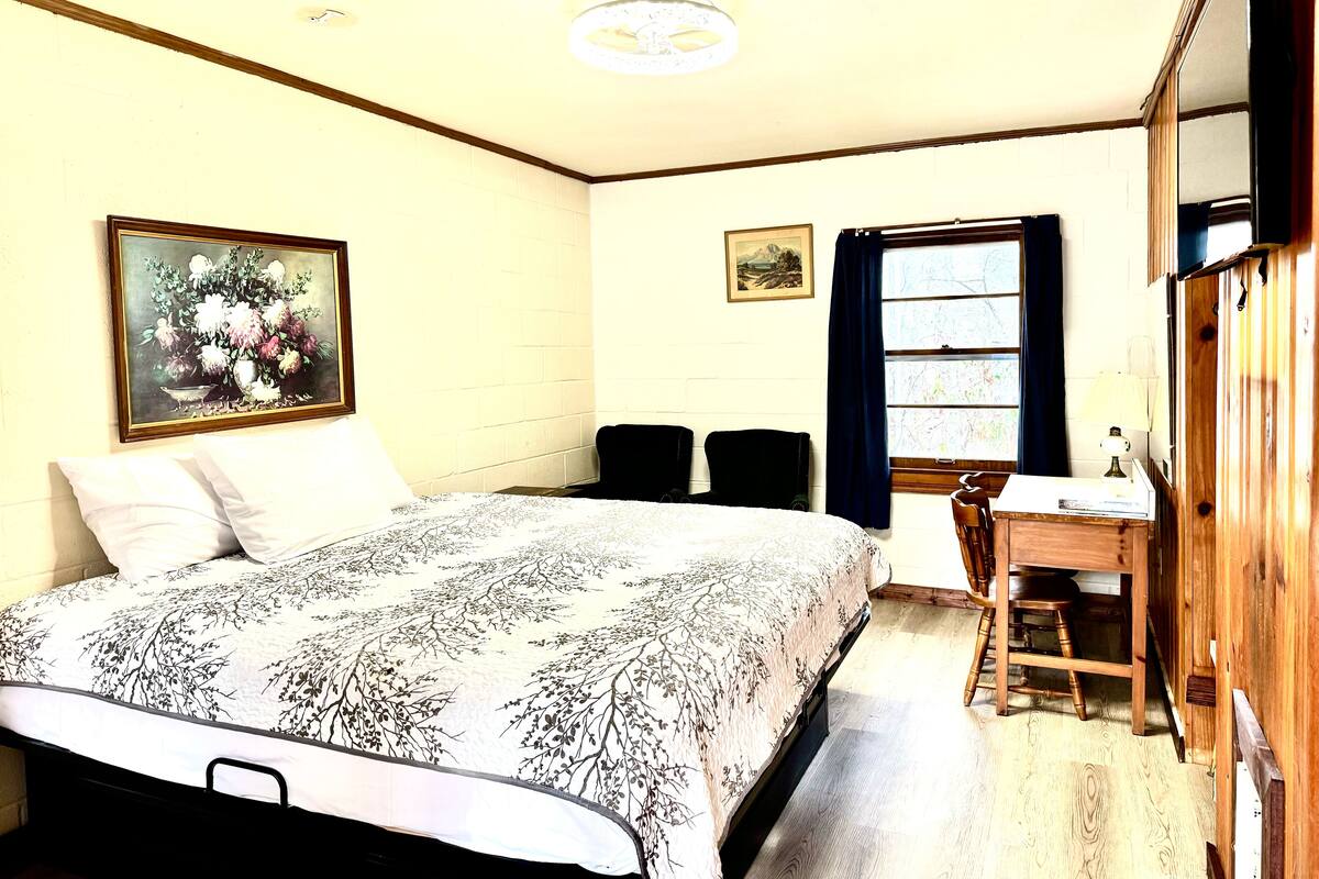 A bedroom with a bed, two chairs, a desk, a window with curtains, and two framed pictures on the wall.
