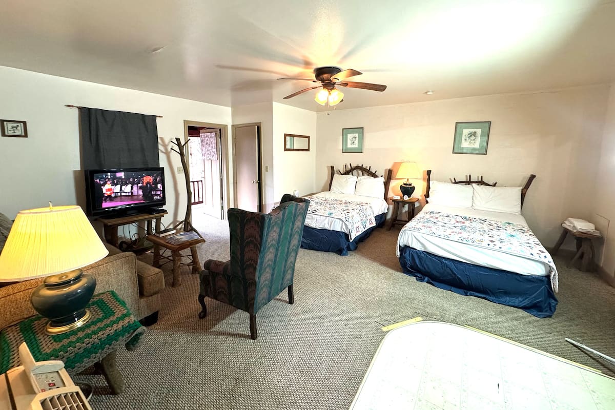 A hotel room with two beds, a television, a ceiling fan, and a small table with a lamp.