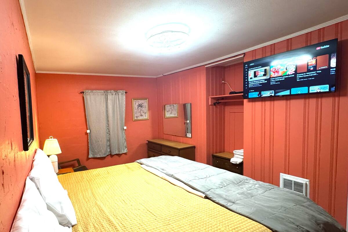 A bedroom with a bed, a television mounted on the wall, a window with curtains, and a small table with a lamp.